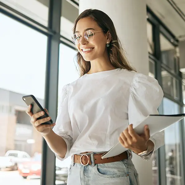 Lächelnde Frau mit Smartphone und Tablet läuft durch ein modernes Bürogebäude.