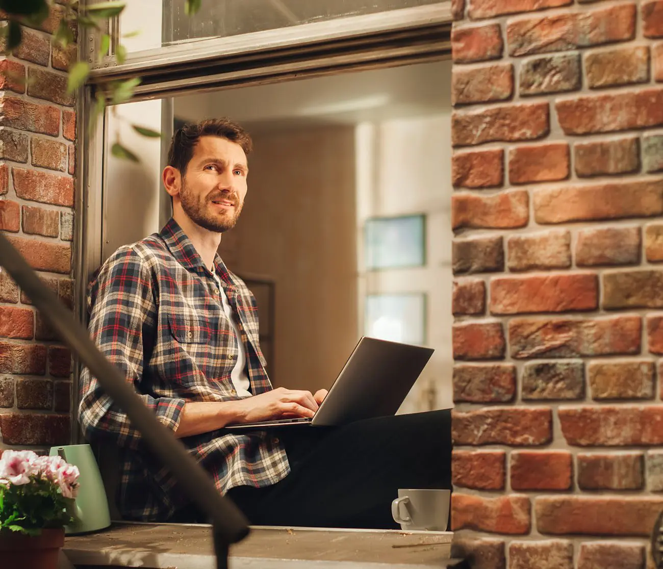 Mann sitzt auf dem Fensterbrett mit Laptop auf dem Schoß und denkt über UI/UX-Design nach